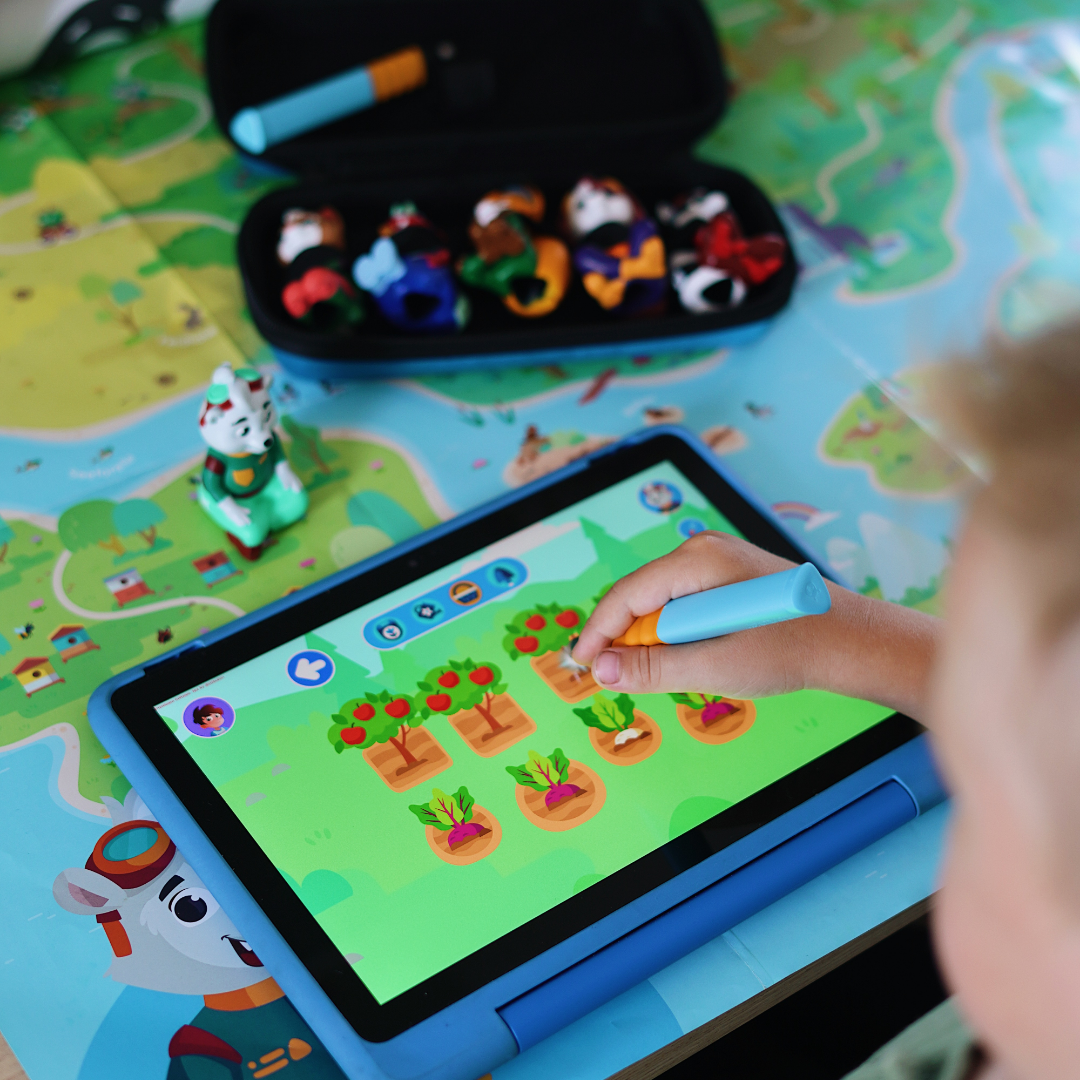 Ein Kind spielt mit einem Stift ein Bauernhofspiel auf einem Tablet, im Hintergrund ist das Angebot Entdecker-Bundle mit Spielzeug und einer bunten Karte zu sehen.